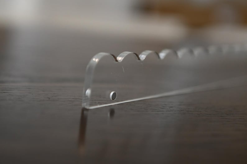 May include: A clear acrylic cake server with a scalloped edge. The server has a flat, rectangular shape with a series of rounded scallops along one side. The server is designed for cutting and serving cake.