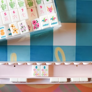 May include: A complete Mahjong set with white tiles featuring colorful symbols and characters, including flowers, bamboo, and dragons. The set includes a clear plastic case and a pink tile rack, all set against a blue and white checkered background.