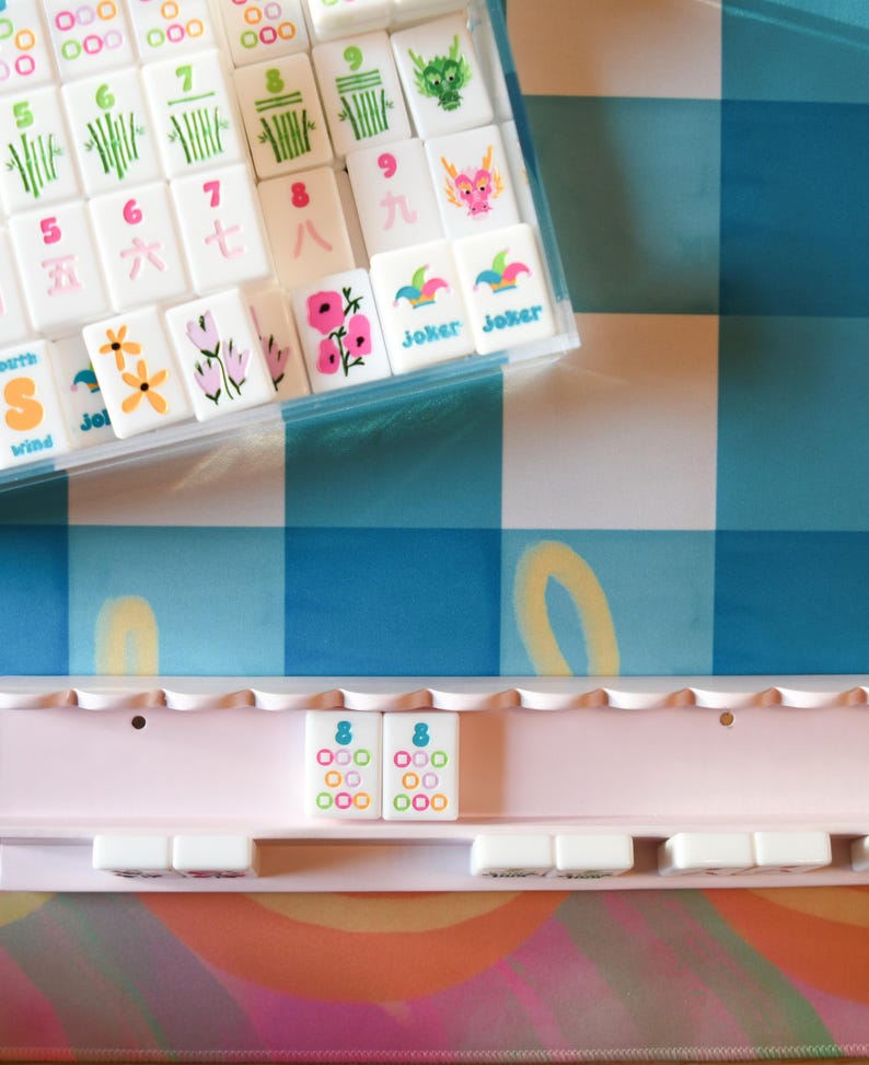 May include: A complete Mahjong set with white tiles featuring colorful symbols and characters, including flowers, bamboo, and dragons. The set includes a clear plastic case and a pink tile rack, all set against a blue and white checkered background.