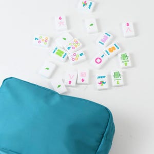 May include: A teal zippered pouch accompanied by a collection of white rectangular Mahjong tiles. The tiles feature colorful printed designs including numbers, bamboo stalks, wind directions, and joker icons in pink, green, blue, and orange.