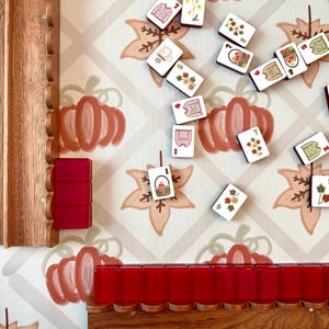 May include: A Mahjong game set featuring white tiles with colorful autumn-themed illustrations of sweaters, baskets, and leaves, arranged on a patterned surface with pumpkins and maple leaves, alongside wooden racks and solid red tiles.