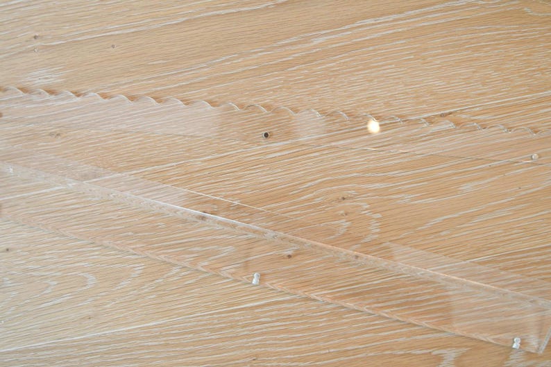 May include: Close-up of a light-colored wooden surface with visible grain and a clear, angled overlay. The overlay appears to be made of a transparent material, possibly acrylic or glass, and is secured with small fasteners.