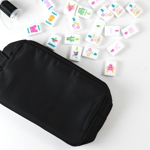 May include: A solid black rectangular zippered pouch positioned next to a collection of white Mahjong tiles featuring colorful symbols and numbers, along with a small spool of black thread, all arranged on a plain white surface.