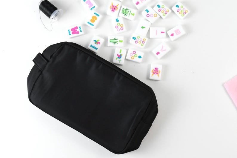 May include: A solid black rectangular zippered pouch positioned next to a collection of white Mahjong tiles featuring colorful symbols and numbers, along with a small spool of black thread, all arranged on a plain white surface.