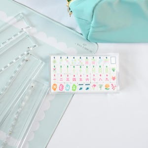 May include: A rectangular clear acrylic case containing a set of mahjong tiles with colorful printed designs, including numbers one through nine, Chinese characters, floral motifs, and the word NEWS, alongside a light blue zippered pouch.