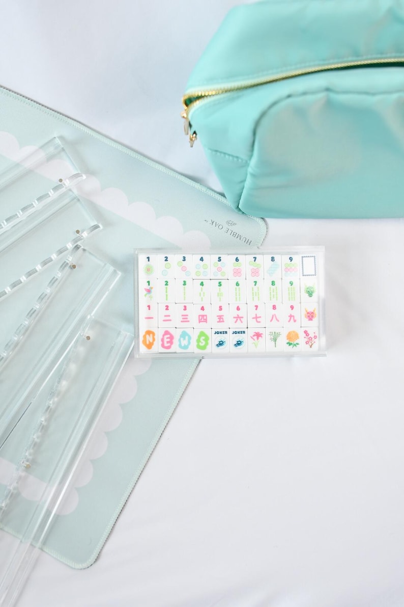 May include: A rectangular clear acrylic case containing a set of mahjong tiles with colorful printed designs, including numbers one through nine, Chinese characters, floral motifs, and the word NEWS, alongside a light blue zippered pouch.