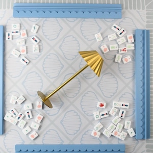 May include: A board game set featuring a light blue playing surface with a white seashell pattern, four light blue tile racks, and numerous white rectangular tiles with colorful icons. A gold-colored metal lamp stands in the center.