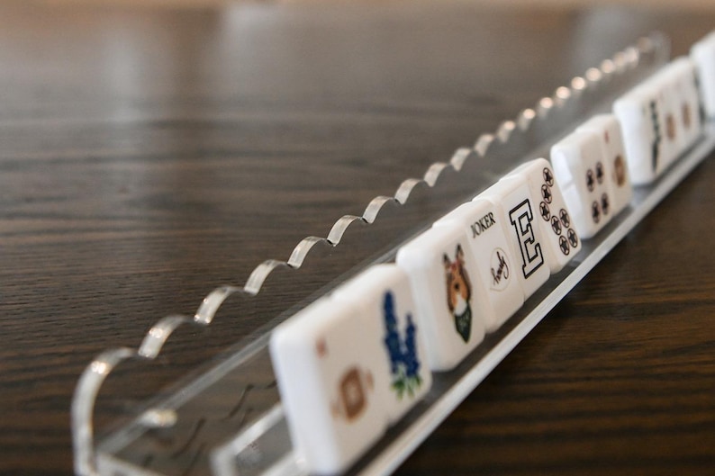 May include: A clear acrylic domino holder with a wavy edge, holding several white dominoes. The dominoes have various designs, including a blue flower, a dog, the letter "E", and other symbols. The holder sits on a dark wooden surface.