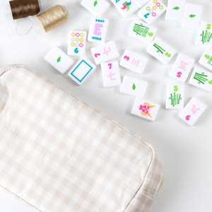 May include: A beige and white gingham fabric pouch positioned next to a collection of white mahjong tiles featuring colorful symbols, numbers, and the word "Joker" or "East Wind," along with two small spools of brown and gold sewing thread.