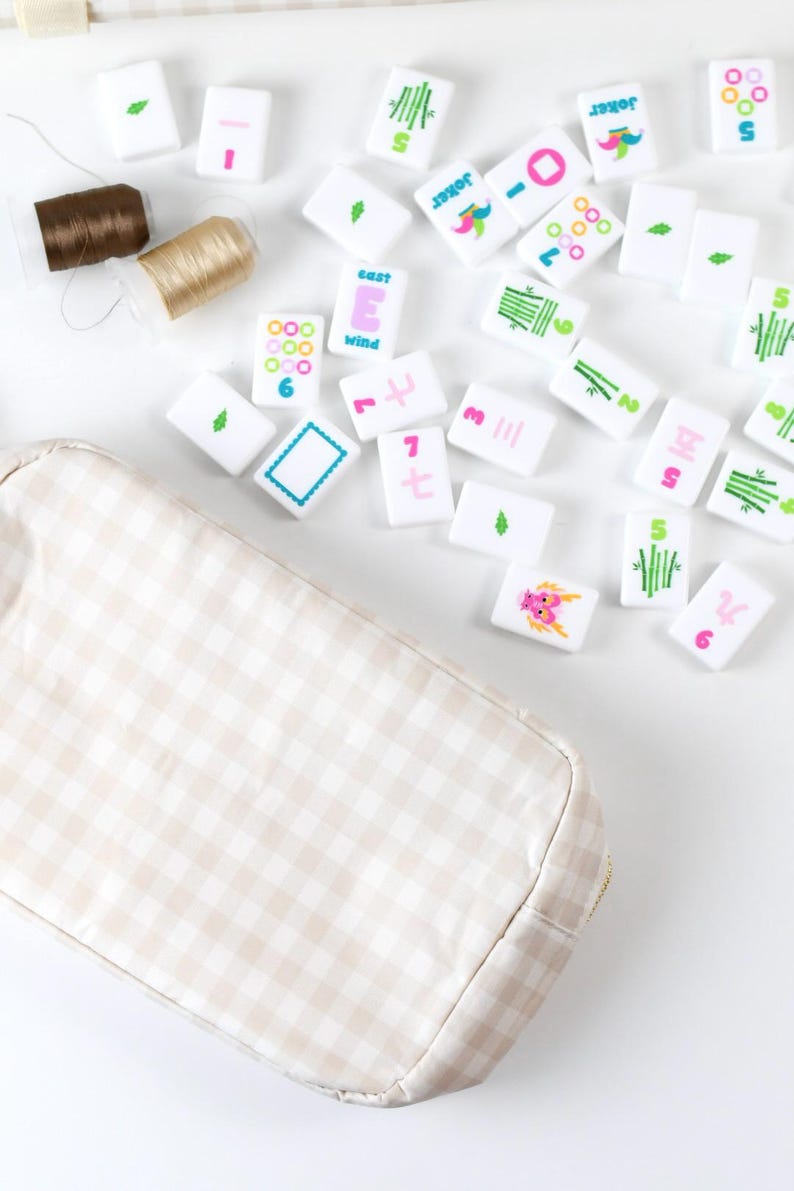 May include: A beige and white gingham fabric pouch positioned next to a collection of white mahjong tiles featuring colorful symbols, numbers, and the word "Joker" or "East Wind," along with two small spools of brown and gold sewing thread.