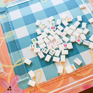 May include: A collection of white Mahjong tiles scattered on a blue and white checkered game board. Some tiles feature colorful symbols and letters. The board is set on a floral patterned surface.