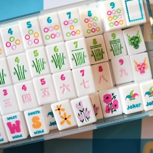 May include: A set of white Mahjong tiles with colorful symbols and numbers, including bamboo stalks, flowers, and dragons. The tiles are arranged in a clear plastic case, set against a blue and white checkered background.