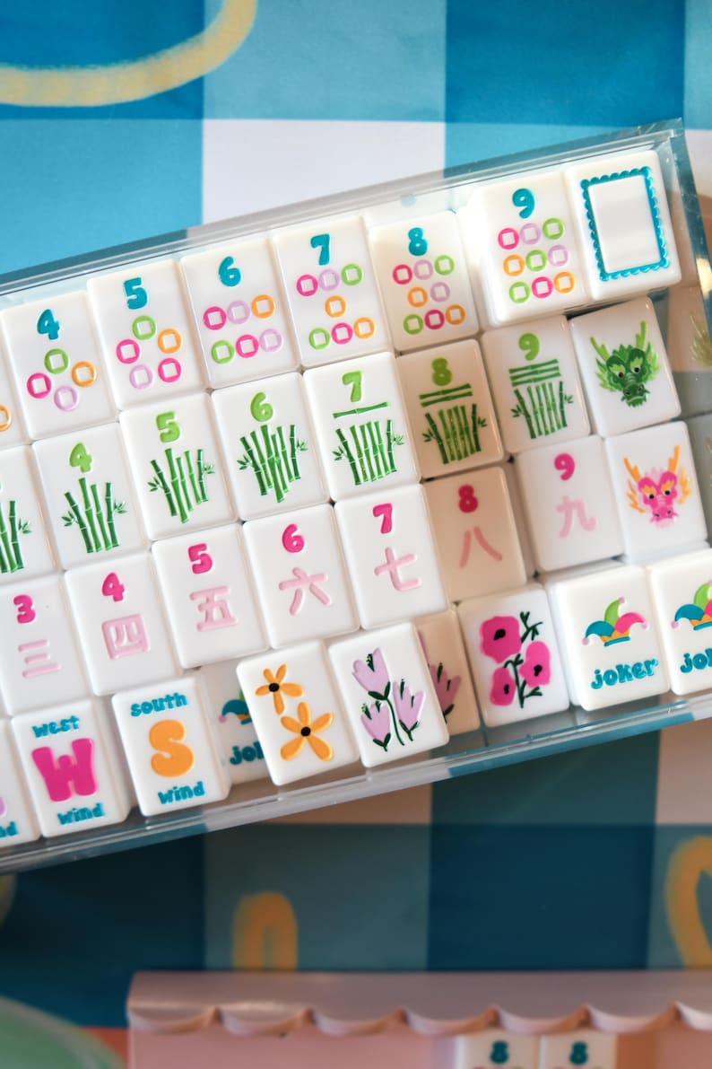 May include: A set of white Mahjong tiles with colorful symbols and numbers, including bamboo stalks, flowers, and dragons. The tiles are arranged in a clear plastic case, set against a blue and white checkered background.