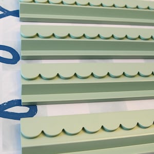 May include: Four sage green decorative wooden trim pieces with a scalloped edge. The trim pieces are arranged in parallel, with a patterned background of blue swirls and a checkered pattern.