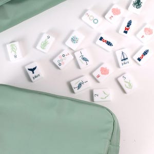 May include: A set of white rectangular mahjong tiles featuring various nautical and nature-themed illustrations, including a sailboat, lighthouse, octopus, and heron, alongside numbers and the word joker, stored in a sage green fabric carrying case.