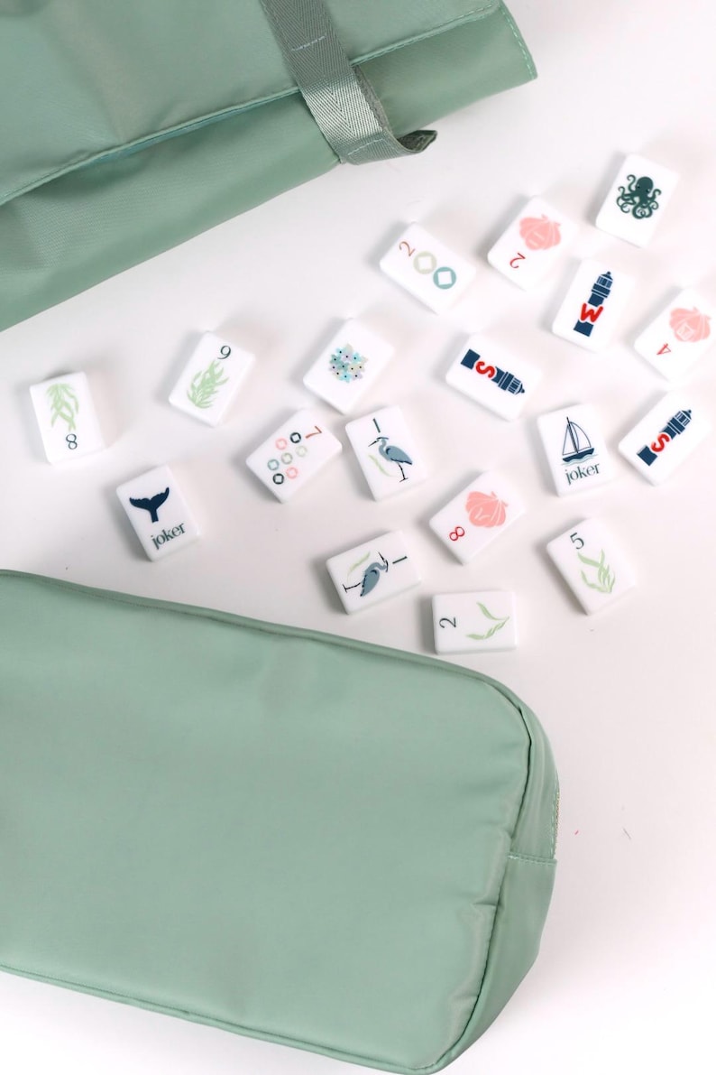 May include: A set of white rectangular mahjong tiles featuring various nautical and nature-themed illustrations, including a sailboat, lighthouse, octopus, and heron, alongside numbers and the word joker, stored in a sage green fabric carrying case.