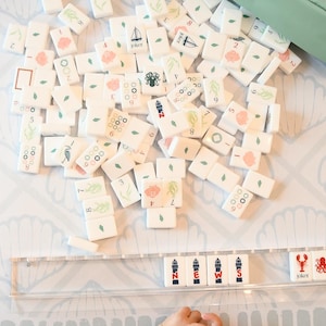May include: A collection of white mahjong tiles with nautical-themed illustrations including seashells, lighthouses, octopuses, and lobsters. A clear acrylic rack displays tiles spelling the word NEWS alongside lobster and octopus icons.