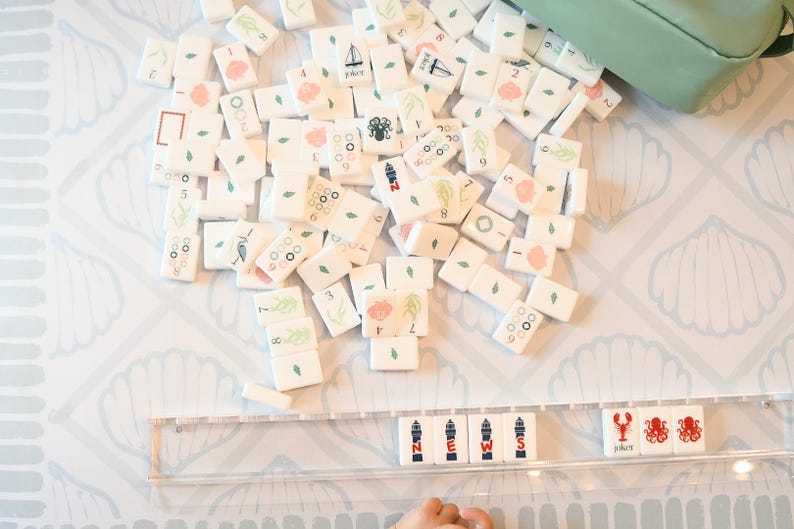 May include: A collection of white mahjong tiles with nautical-themed illustrations including seashells, lighthouses, octopuses, and lobsters. A clear acrylic rack displays tiles spelling the word NEWS alongside lobster and octopus icons.