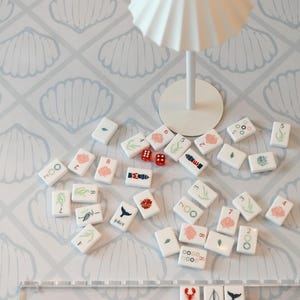 May include: A set of white mahjong tiles featuring colorful illustrations of sea life, numbers, and symbols, including three joker tiles with a lobster, sailboat, and whale. Two small red dice and a white pleated lamp are also visible.