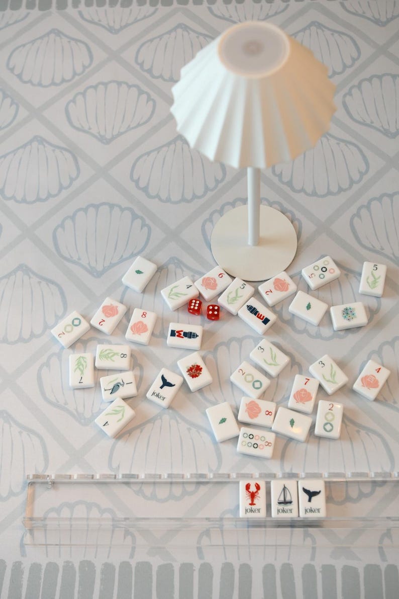 May include: A set of white mahjong tiles featuring colorful illustrations of sea life, numbers, and symbols, including three joker tiles with a lobster, sailboat, and whale. Two small red dice and a white pleated lamp are also visible.