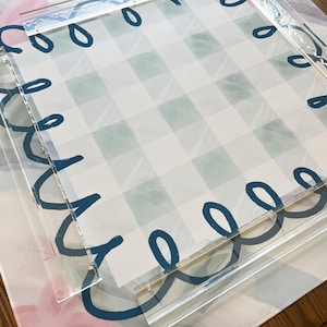 May include: A clear acrylic tray with a teal loop design around the edges, layered over a patterned surface. The surface features a blue and white checkered pattern and a floral design in pink and blue.