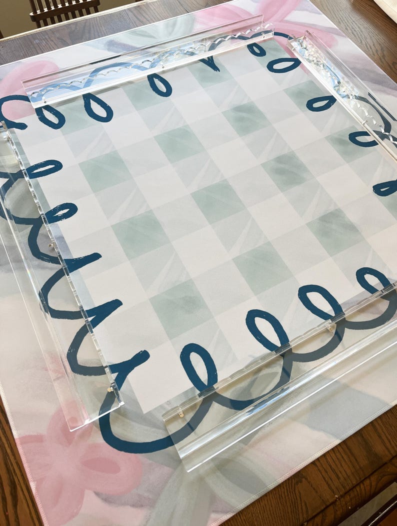 May include: A clear acrylic tray with a teal loop design around the edges, layered over a patterned surface. The surface features a blue and white checkered pattern and a floral design in pink and blue.