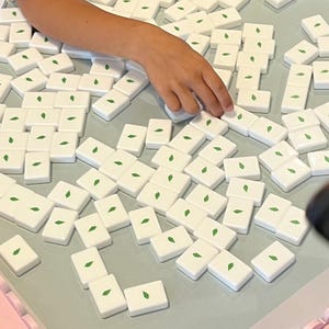 May include: A close-up shot of a board game with white tiles featuring a green leaf design. The tiles are scattered on a light blue surface, with pink decorative borders. A hand is reaching for a tile.