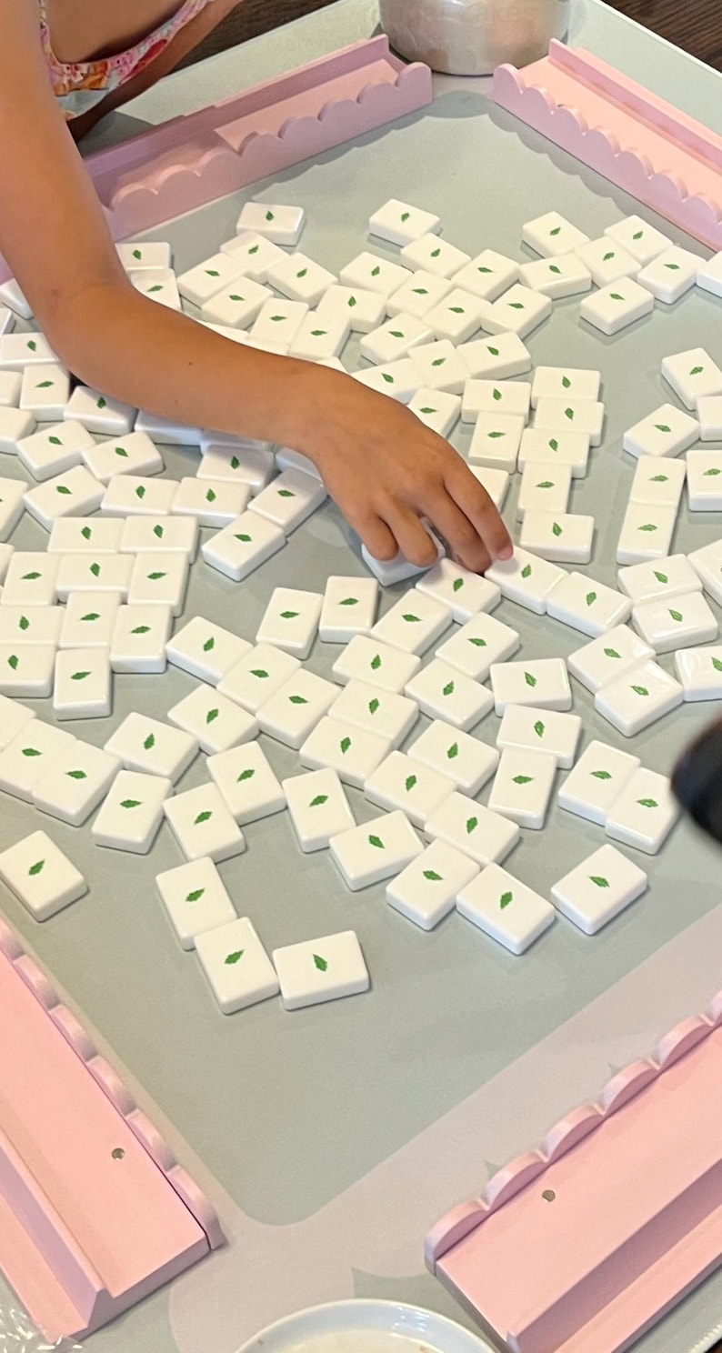 May include: A close-up shot of a board game with white tiles featuring a green leaf design. The tiles are scattered on a light blue surface, with pink decorative borders. A hand is reaching for a tile.