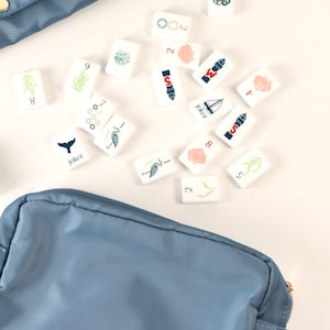 May include: A set of white rectangular game tiles featuring various colorful illustrations including an octopus, a sailboat, a whale tail, and the word Joker, scattered between two blue zippered storage pouches made of smooth fabric.