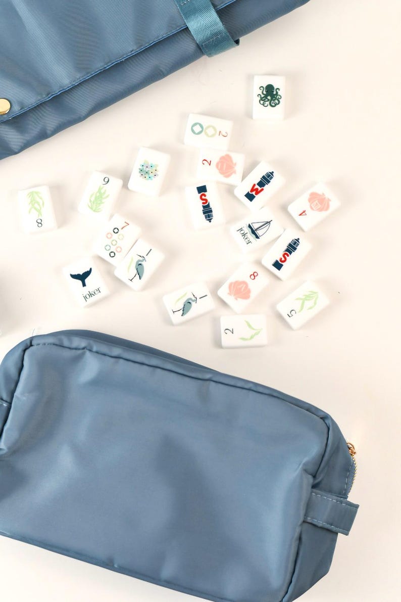 May include: A set of white rectangular game tiles featuring various colorful illustrations including an octopus, a sailboat, a whale tail, and the word Joker, scattered between two blue zippered storage pouches made of smooth fabric.