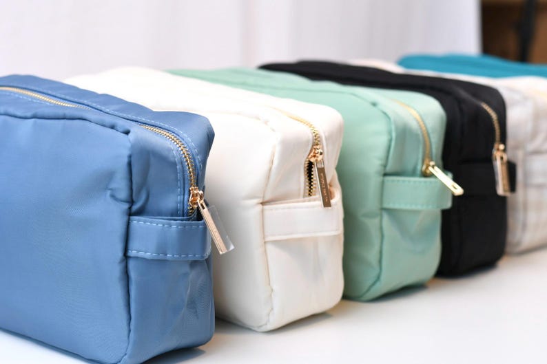 May include: A row of rectangular zippered pouches in various solid colors including blue, white, mint green, and black. Each pouch features a smooth fabric exterior, a gold-toned metal zipper closure, and a small side handle for easy carrying.