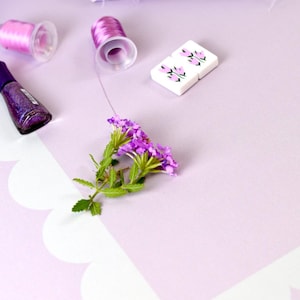 May include: A still life arrangement featuring sewing notions and floral elements. Includes spools of pink thread, a bottle of purple nail polish, fabric, and small white tiles with floral designs. A sprig of purple flowers adds a natural touch.