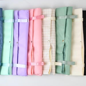 Op de afbeelding: Een collectie van acht rechthoekige stoffen organizers die naast elkaar in een rij zijn geplaatst. De organizers hebben verschillende effen kleuren, waaronder blauw, mint, lavendel, roze, salie, crème en zwart, plus één met een beige en wit ruitpatroon.