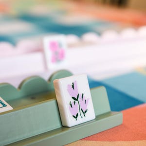 May include: A close-up of a white tile with a pink floral design, resting in a light green wooden holder. The tile features three stylized pink flowers with green stems and leaves. The background includes a colorful patterned surface.