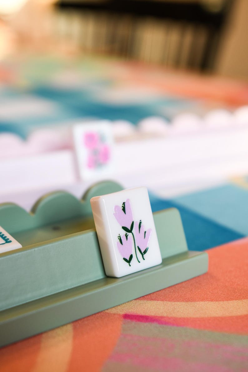May include: A close-up of a white tile with a pink floral design, resting in a light green wooden holder. The tile features three stylized pink flowers with green stems and leaves. The background includes a colorful patterned surface.