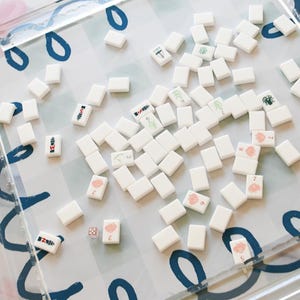 May include: A clear acrylic Mahjong game set with white tiles scattered on a patterned surface. The tiles feature various symbols and numbers in red, green, and black. The game set includes dice and is designed for strategic gameplay.