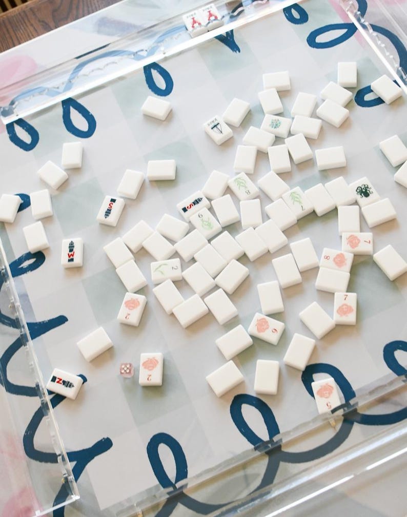 May include: A clear acrylic Mahjong game set with white tiles scattered on a patterned surface. The tiles feature various symbols and numbers in red, green, and black. The game set includes dice and is designed for strategic gameplay.