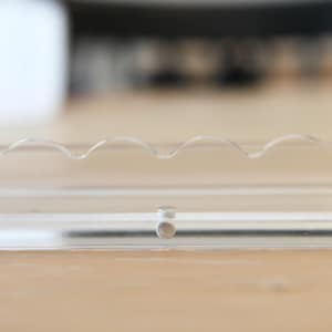 May include: Clear, transparent plastic object with a wavy design on top. The object is rectangular with a small, circular piece in the middle. The background is a light brown wooden surface.