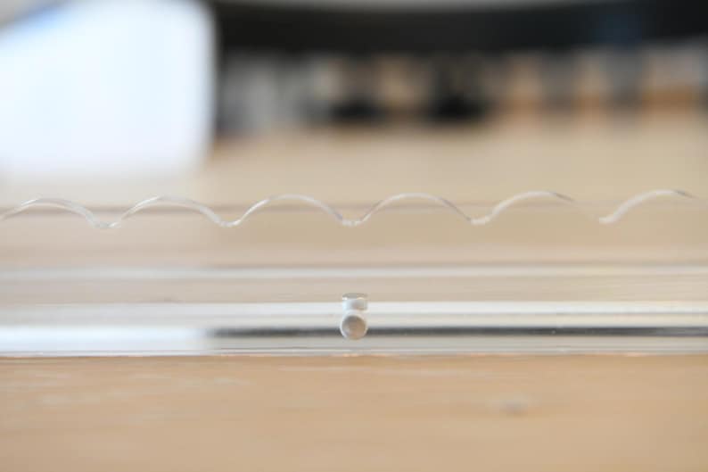 May include: Clear, transparent plastic object with a wavy design on top. The object is rectangular with a small, circular piece in the middle. The background is a light brown wooden surface.