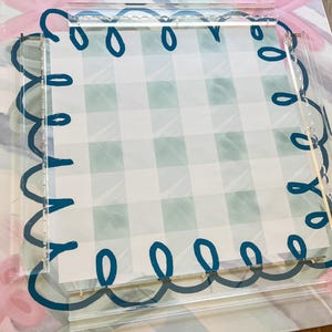 May include: A clear acrylic tray with a scalloped edge design in dark teal. The tray frames a light green and white checkered pattern. The background features a floral and bow design in pink and blue.