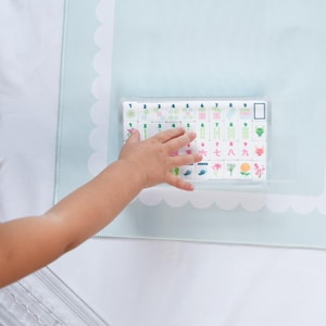 May include: A set of white rectangular mahjong tiles arranged in a clear plastic tray, featuring colorful printed numbers, symbols, and floral designs in rows, with a light blue scalloped border mat visible underneath the tray.