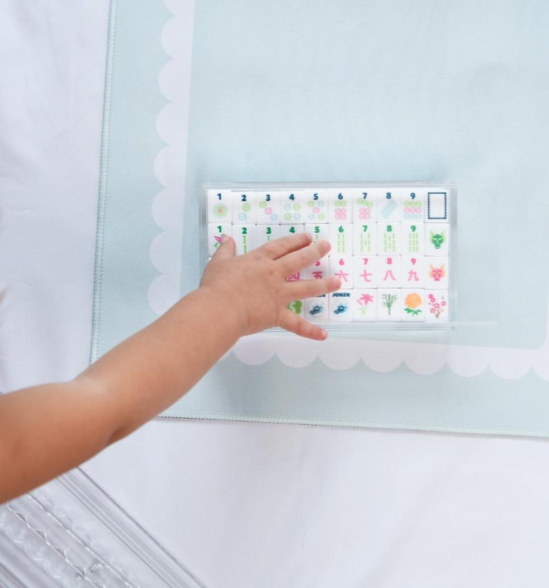 May include: A set of white rectangular mahjong tiles arranged in a clear plastic tray, featuring colorful printed numbers, symbols, and floral designs in rows, with a light blue scalloped border mat visible underneath the tray.