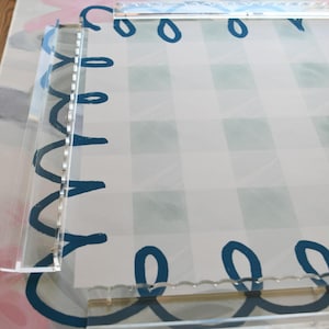 May include: Clear acrylic frame with a decorative border of blue loops and a gingham pattern background. The frame is designed to hold a photo or artwork. The background has a floral design.