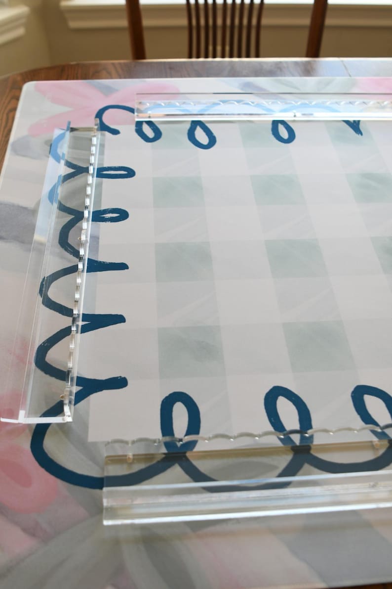 May include: Clear acrylic frame with a decorative border of blue loops and a gingham pattern background. The frame is designed to hold a photo or artwork. The background has a floral design.