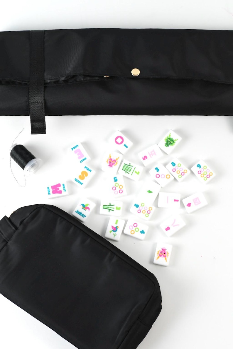 May include: A set of white rectangular mahjong tiles featuring colorful printed symbols, numbers, and characters, accompanied by a black fabric carrying case, a black fabric storage roll with a gold snap button, and a small spool of black thread.