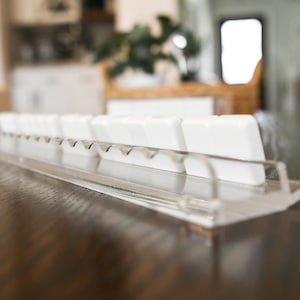 May include: A clear acrylic keyboard key holder with a row of white keycaps. The holder has a clear, angled base and a series of small notches to hold the keycaps in place. The background is blurred, showing a wooden surface.