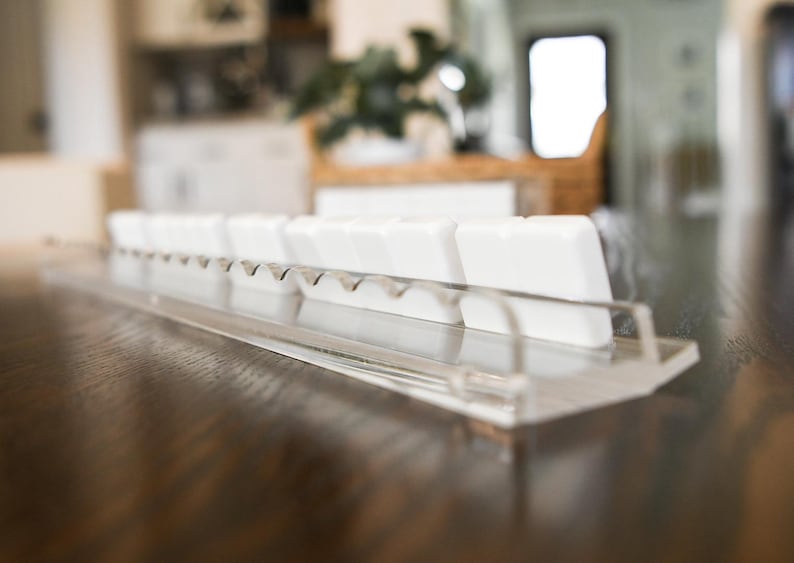 May include: A clear acrylic keyboard key holder with a row of white keycaps. The holder has a clear, angled base and a series of small notches to hold the keycaps in place. The background is blurred, showing a wooden surface.