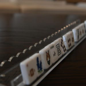 May include: A clear acrylic tile holder with a scalloped edge, holding several white tiles. The tiles have various symbols and images, including a horse, a flower, and the word "Joker". The holder sits on a dark wooden surface.