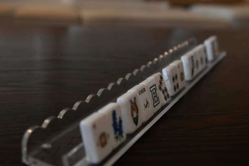 May include: A clear acrylic tile holder with a scalloped edge, holding several white tiles. The tiles have various symbols and images, including a horse, a flower, and the word "Joker". The holder sits on a dark wooden surface.