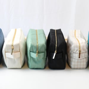 May include: A row of six rectangular zippered pouches in various colors including blue, white, mint green, black, beige gingham, and teal. Each pouch features a gold-toned metal zipper closure and a small fabric side handle for easy carrying.